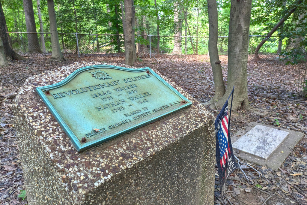A stone pedestal with a green metal plaque that reads: Revolutionary Soldiers; Isaac Walker, 1721 - (illegible); Nathan Walker, 1736 - (illegible); Placed by Prince George's County Chapter D.A.R." Next to it is a drooping black banner with a stars-and-stripes image in blue, white and red.