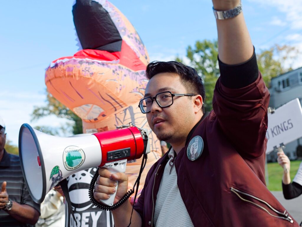 Fritz, a light-brown-skinned man with short dark hair and glasses, holds a red and white megaphone in his right hand with his left hand upraised. He wears a white shirt and red jacket. Behind him stands someone in an orange inflatable dinosaur costume.