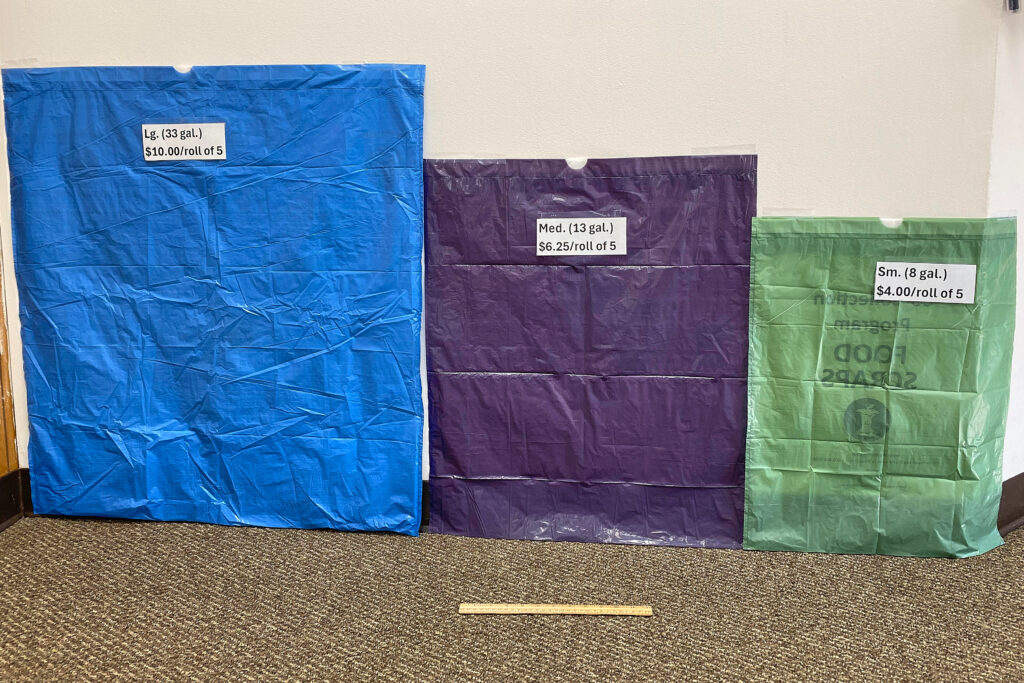 Three trash bags - large blue, medium purple and small green - are pinned up flat against a wall. A ruler is on the floor in front of the medium bag, which is wider than the ruler.