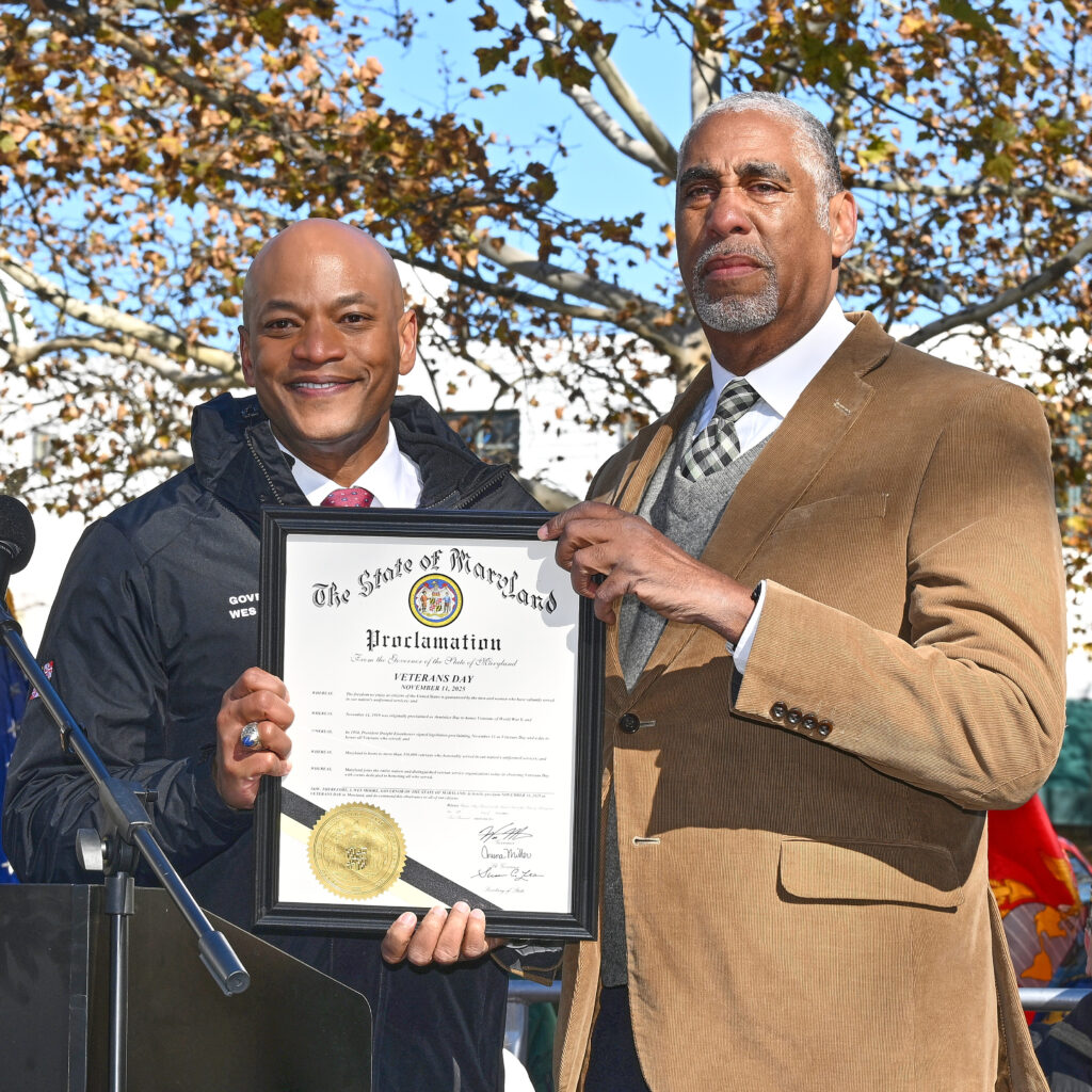 At left, Moore, a brown-skinned man in a black jacket, smiles and holds a proclamation in a frame. At right, Jordan (a brown-skinned man in a tan jacket) places one hand on the proclamation's upper corner, with a somber expression. Behind their heads, tree branches spread against a white building and a blue sky.