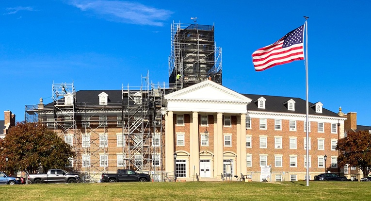 A beige building with many windows, a dark roof, and four white columns supporting a triangular roof structure in the center. One side of the building is surrounded by dark scaffolding. In front of the building, there's a green lawn and a flagpole flying the USA flag.
