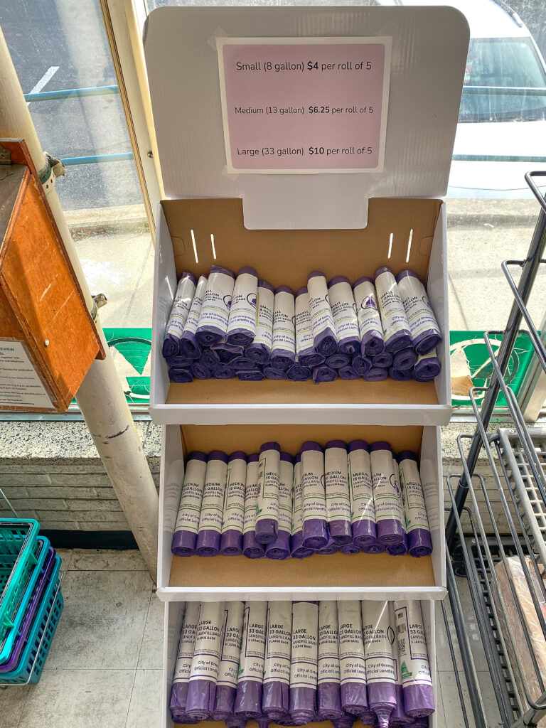 A 3-tiered cardboard rack holding rows of purple bag bundle rolls, each wrapped in a white label. A purple sign at the top of the rack gives the prices.