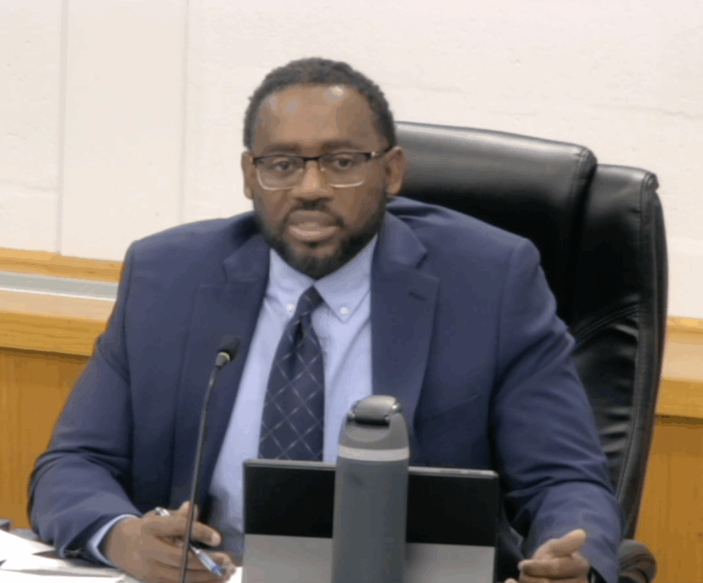 A brown-skinned man with short black hair, mustache and beard. He wears a blue suit and sits in a black office chair. A tablet and a reusable water bottle are on the table in front of him.