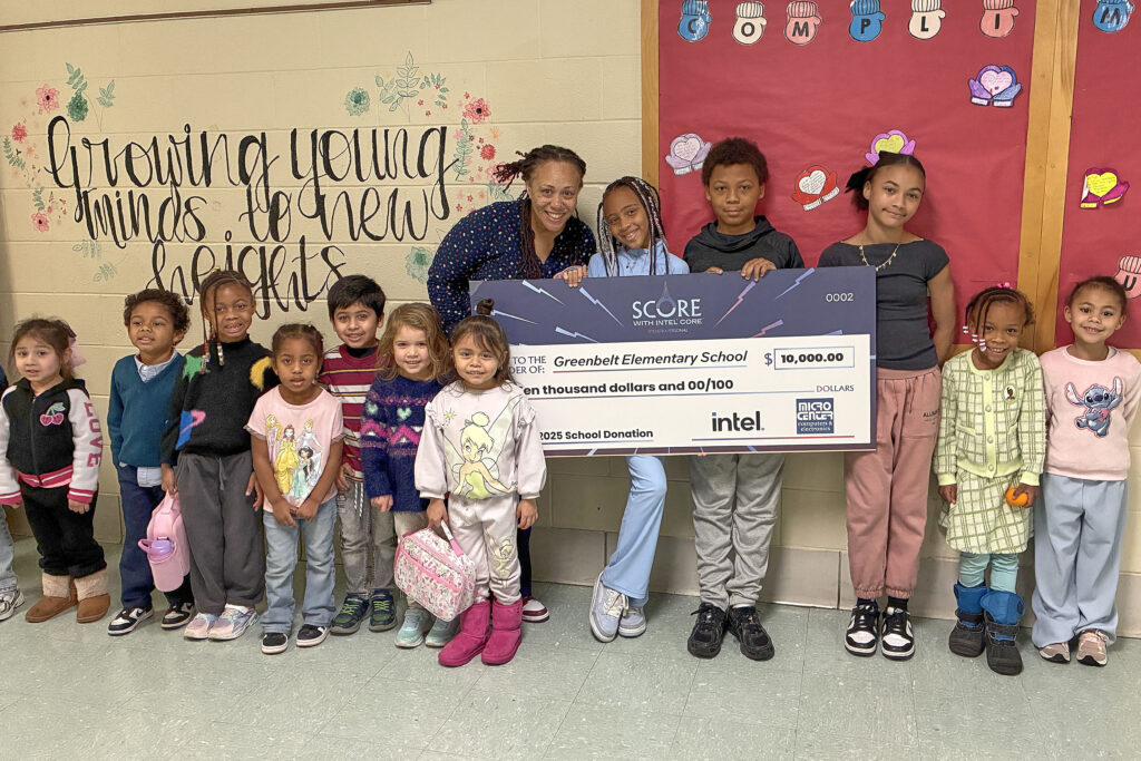 A woman bends down to hold a large display check alongside three older-elementary students. At left are a group of early-elementary students. At right are two middle-elementary students. Behind the group is a painted mural with flowers and the text "Growing young minds to new heights" in swirly letters. At right is a red bulletin board with colorful mitten-and-heart shapes.