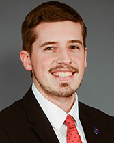 A smiling pale-skinned man with short brown hair and a close-trimmed brown mustache and beard. He wears a white collared shirt, a black jacket, and a red tie, against a gray background.
