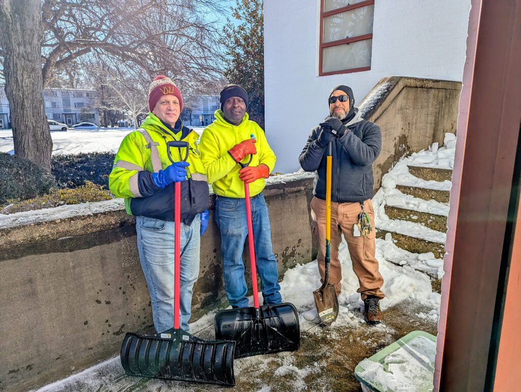 Three men in winter clothes, holding snow shovels, stand in an outdoor stairwell. Snow is mostly cleared from the area where they are standing. Snow is still seen on the steps.
