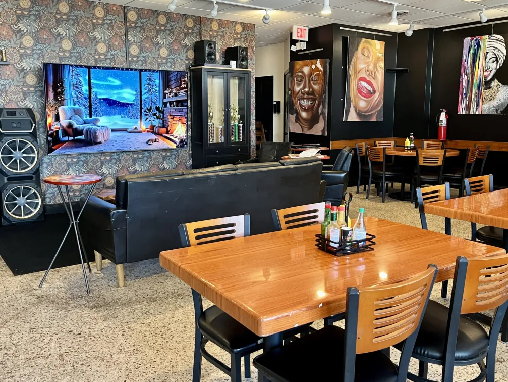 A brown wood restaurant table (4-seat) in the foreground. In the background, couches surround a TV with a wintry scene displaying. Paintings are on the black walls.
