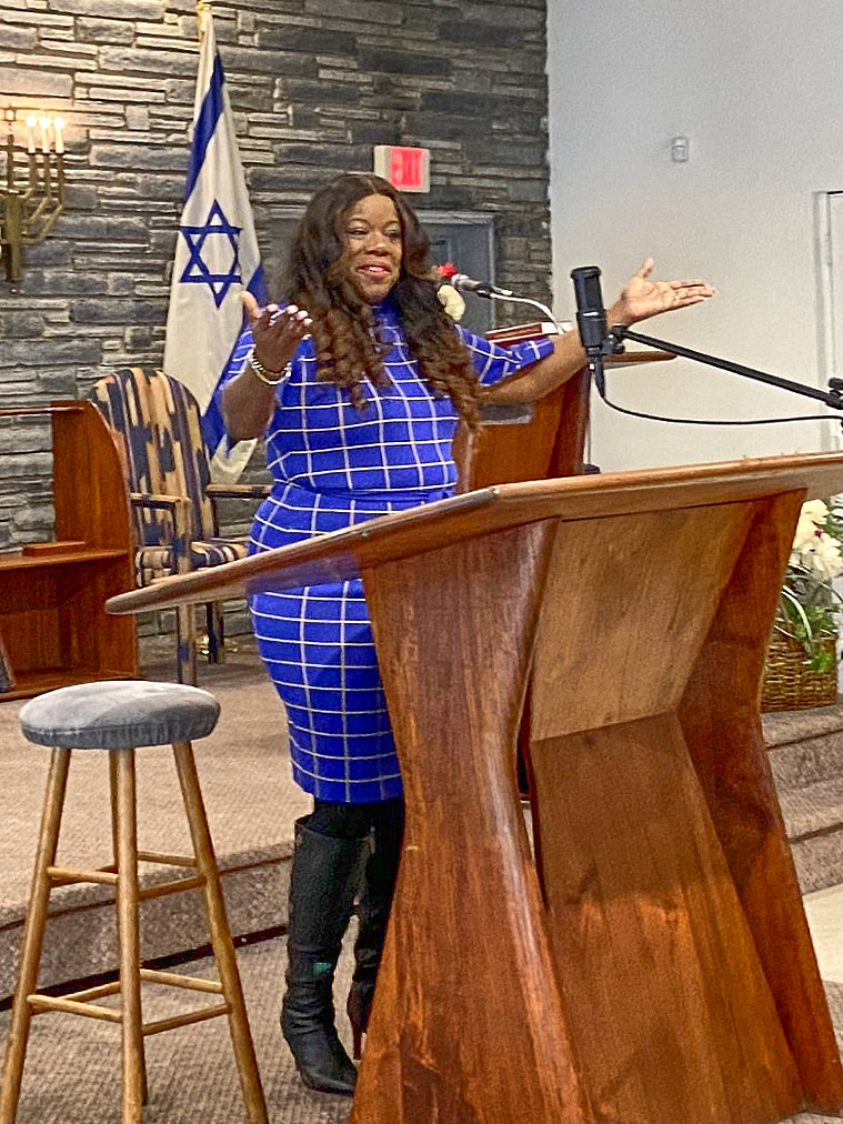 A brown-skinned woman with long brown hair, in a blue dress with a white grid pattern, and black boots. She stands at a wooden lectern with upraised hands and wide-spread arms. Behind her is a flag of Israel draping down from an indoor pole-stand, in front of a gray stone wall.