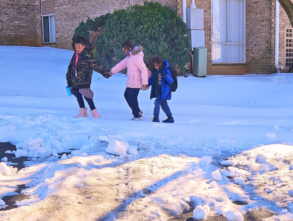 Three children, holding hands, walk across snow, in front of a brown brick apartment building and a green hedge. The three children are in black, pink, and blue winter coats, left to right, respectively.