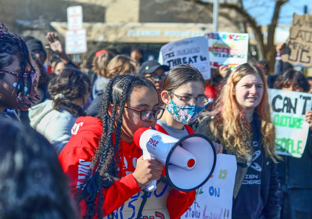 A crowd of high school students. At the front of the crowd, a brown-skinned student with long black braids, in a red sweatshirt and black glasses, holds a red and white megaphone. To her left is a fair-skinned student with brown hair pulled back, glasses, and a colorful face mask. To their left is a fair-skinned student with long loose reddish-blond hair.