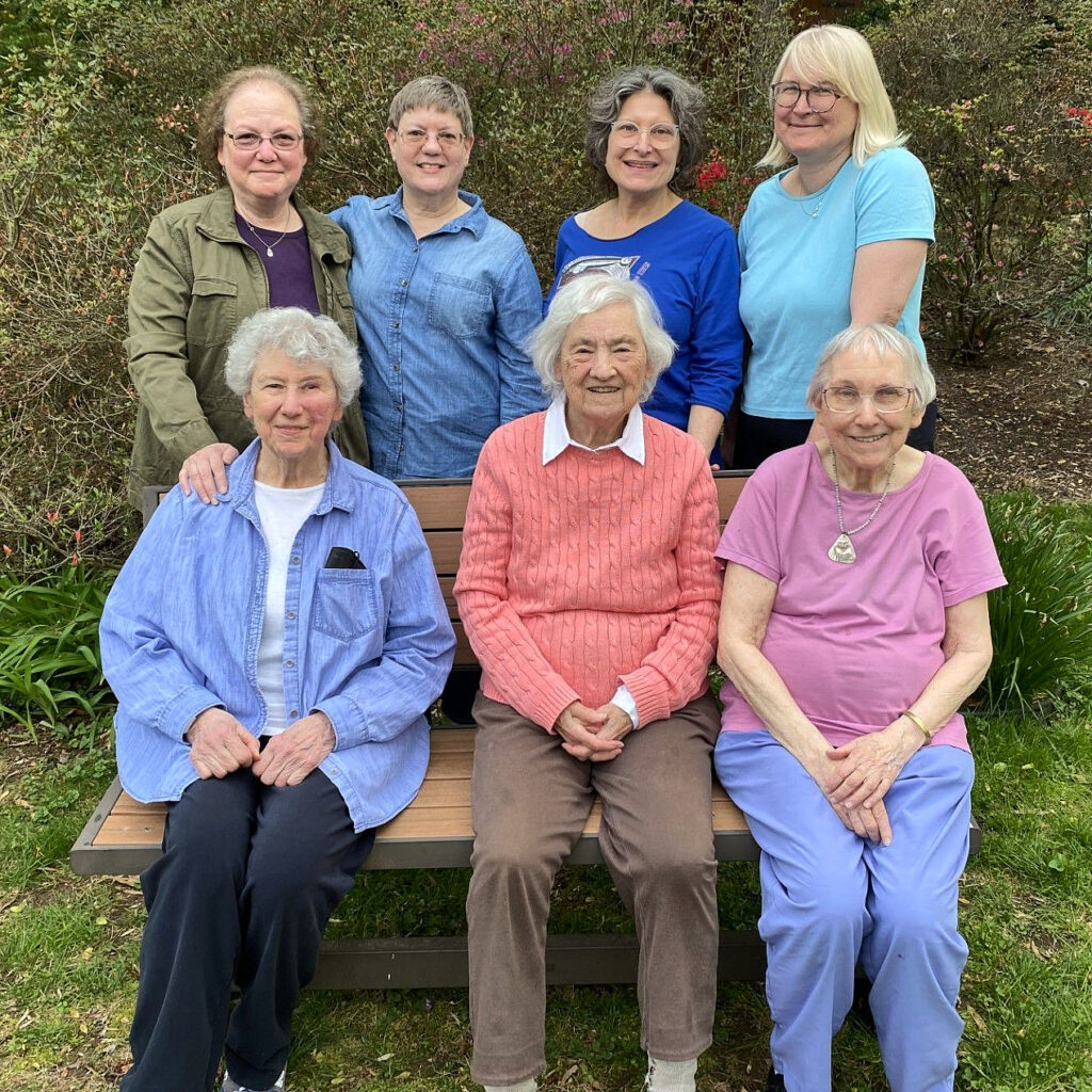 Three white-haired women in colorful clothes sit on an outdoor bench. Behind them stand four women with brown or blond hair.