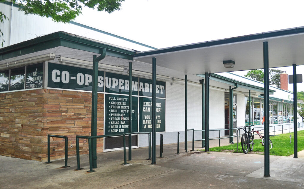 A white flat-roofed building with a green sign reading CO-OP SUPERMARKET and smaller words in white text. One panel of brown brick near the sign. There is a covered walkway outside the building.