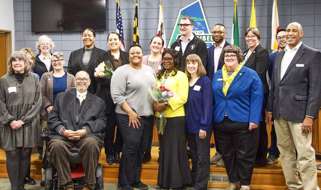 Honorees and local politicians stand together in the Greenbelt City Council chamber, with flags and the Greenbelt logo behind them.