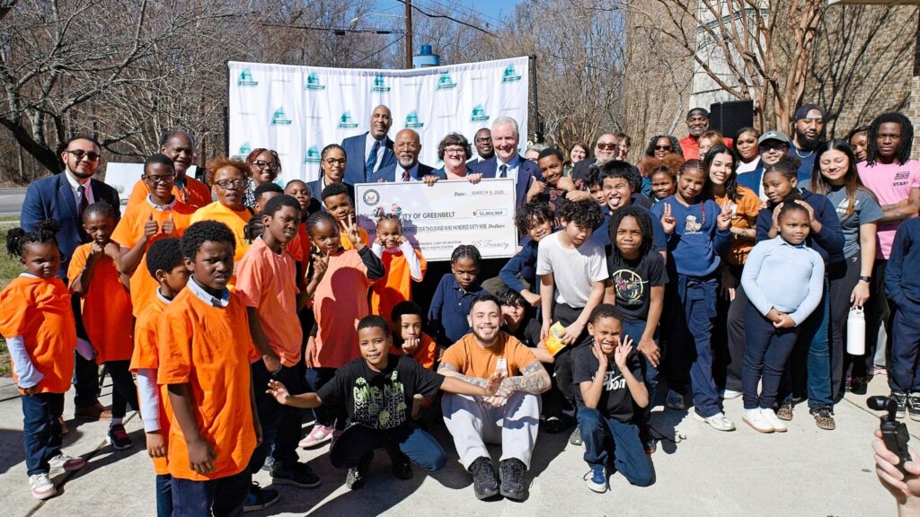 A group of children stand around and in front of adults holding a large presentation check. About half the children wear orange T-shirts, and half wear dark blue T-shirts.