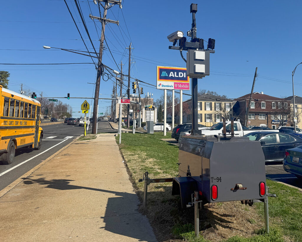 A large black camera with two small red lights at the bottom edge sits on a patch of grass next to a sidewalk. Beside the sidewalk, a school bus drives past. In the background is an Aldi sign.