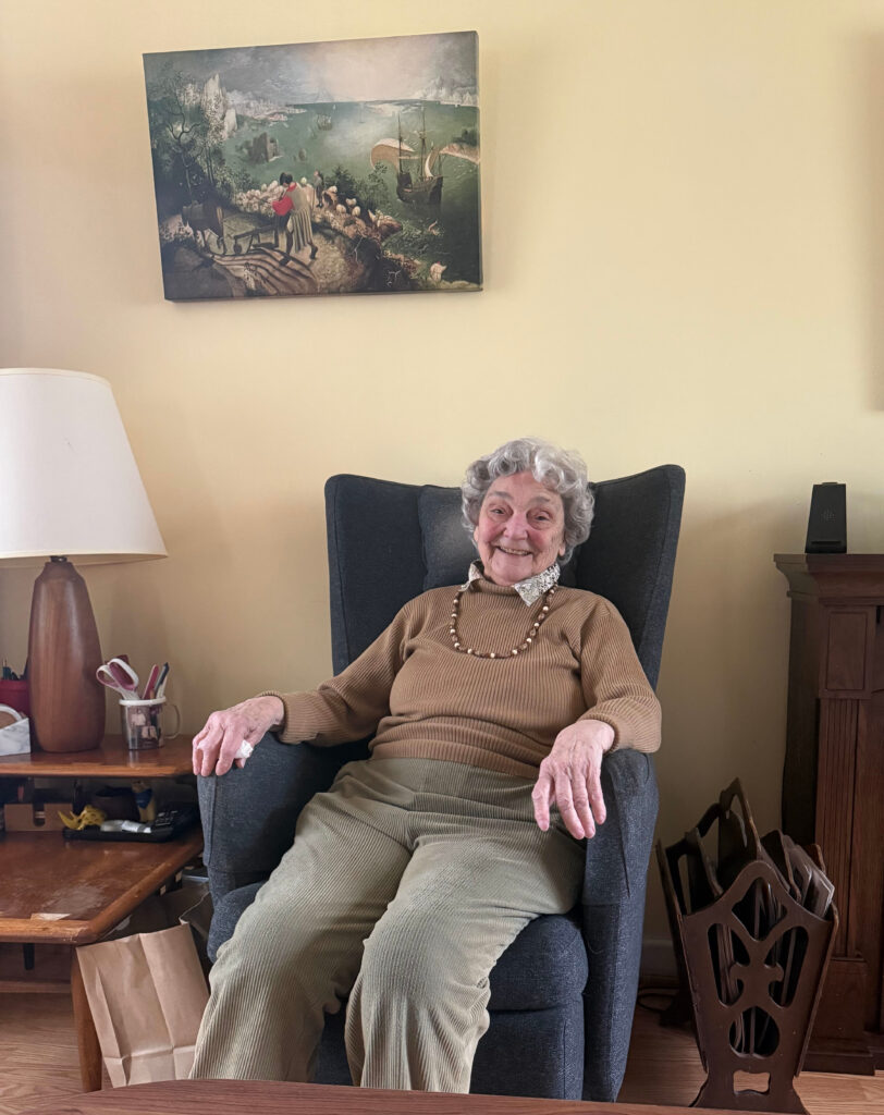 Sylvia Lewis, a light-skinned woman with gray hair, smiles while seated in an armchair in her home.