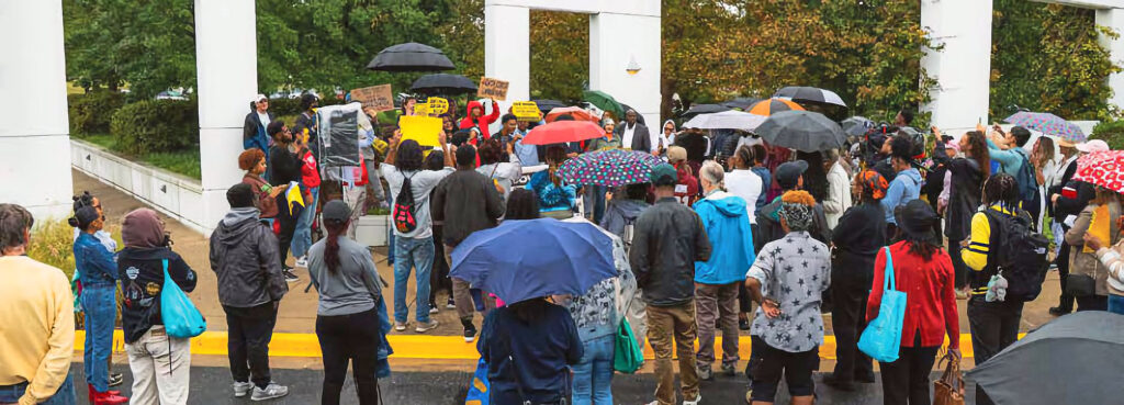 A crowd of people outdoors, on a sidewalk and in the street by the curb. Some hold umbrellas.
