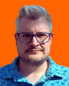 A light-skinned man with short blond hair and glasses, in a blue shirt, with an orange backdrop.