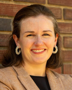 A smiling light-skinned woman with long brown hair and small hoop earrings, wearing a black shirt and brown suit jacket, against a backdrop of a brick wall.