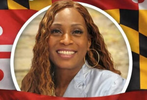 A smiling brown-skinned woman with long curly light brown hair, in a light blue shirt and hoop earrings, with a backdrop of the Maryland flag.