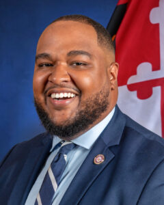 A smiling brown-skinned man with short black hair and beard, wearing a blue suit jacket with round lapel pin, light blue shirt, and blue and white striped tie. The background is blue with a Maryland flag behind him.
