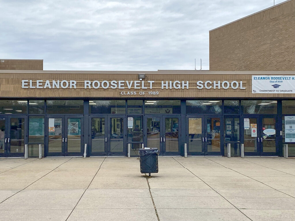 A brown school building with glass doors. The words ELEANOR ROOSEVELT HIGH SCHOOL are in silvery text above the doors.