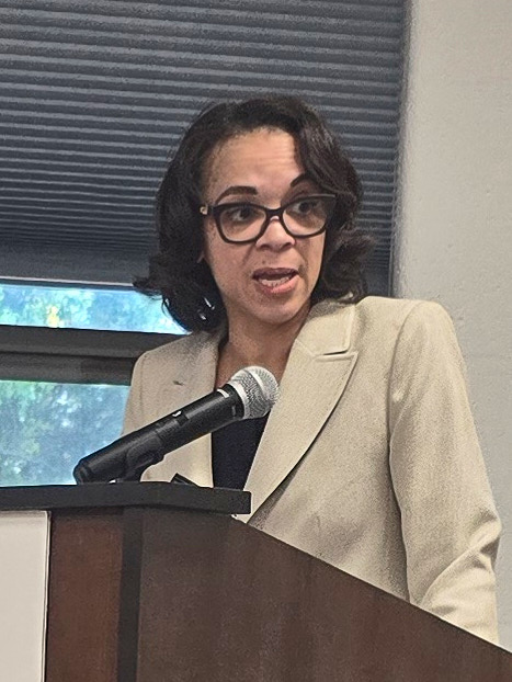 A woman with light tan skin and shoulder-length dark hair speaks into a microphone at a podium. She wears black-rimmed glasses and an ecru suit jacket over a black blouse.