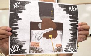 Art piece: An arm in a gray suit, with a brown-skinned fist, pushing downward into a ballot box while holding a paper reading Voting Rights Act of 1965. A yellow rope wraps around the hand's finger, leading downward into the box. Inside the ballot box, shadowy figures hold up pink signs reading "Literacy Tests!" and "Grandfather Clause!" In each of the four corners of the artwork, black shading surrounds the word NO! in white text. 