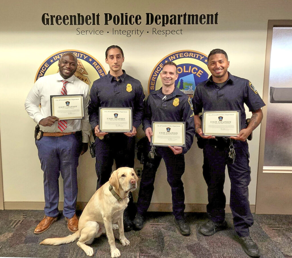 Four police officers stand side-by-side, each holding a framed certificate. A yellow Lab dog sits in front of them. Behind them on the wall are the words Greenbelt Police Department: Service, Integrity, Respect" and two large round logos, mostly obscured by the officers.
