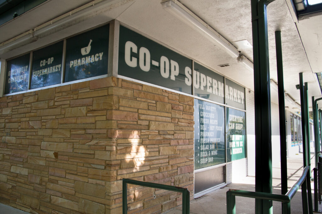 A building with beige stonework and green signage saying "Co-Op Supermarket." and "Pharmacy" in white letters. Metal poles hold up an outdoor roof over a sidewalk corridor.
