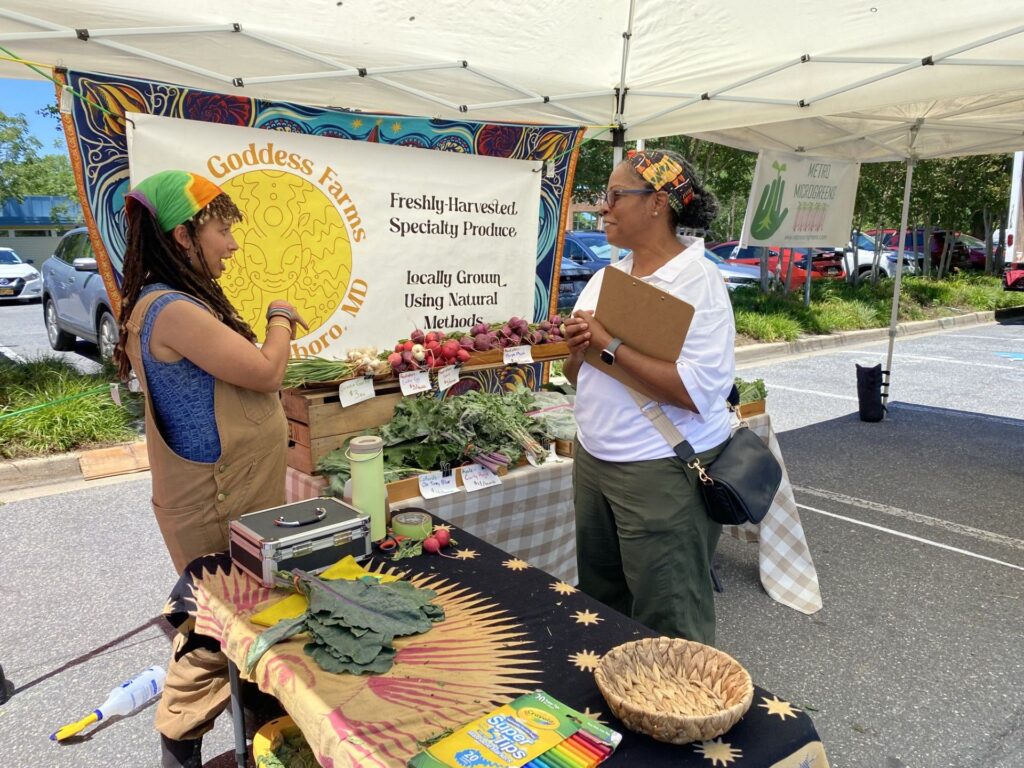 Two people stand, talking, under a white canopy, in front of a "Sun Goddess Farms" sign. The stand's two tables are nearly perpendicular, with vegetables on one and a bright tablecloth on the other.