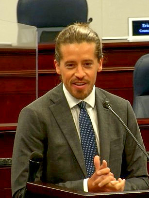 A smiling man with golden-brown hair stands at a podium with a microphone. Behind him is brown wooden seating. He wears a gray suit, white shirt, and blue tie with white dots.