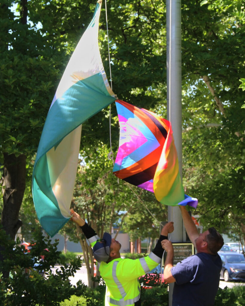 Two workers hold the green-and-white Greenbelt flag and the rainbow-striped Pride flag, while attaching them to the flagpole. The flags billow in the wind.