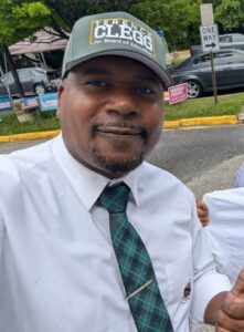 Black man in white shirt and green tie wearing green baseball cap with Terence Clegg on it.
