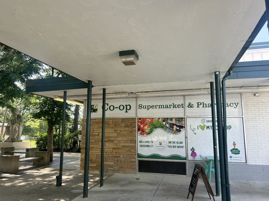 The storefront of Greenbelt Co-op Supermarket and Pharmacy includes poster-covered windows. One poster bears colorful photos of fruit and vegetables with the slogan Welcome to Greenbelt! We're Glad You're Here! Another poster provides a selfie-taking spot: it bears the slogan I Love My Co-Op (with a heart instead of the word Love) overhead, with two cartoon vegetables on the sides, framing a white space to stand in front of.