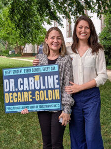 Two women with shoulder-length hair stand outside. One of them holds a blue and yellow campaign poster.