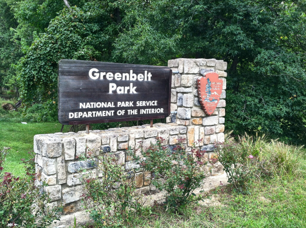 The Greenbelt Park sign, on a stone-wall pedestal with the Greenbelt logo, on grass, in front of trees.