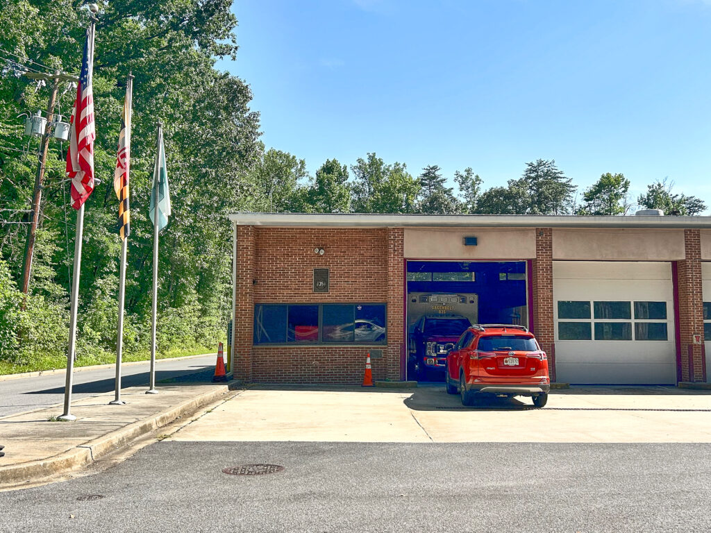 A red car is parked in front of an open vehicle bay in the brown fire department building. A line of flagpoles stands to the left of the building.