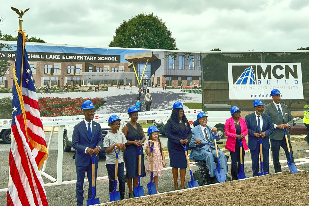 A line of people (including two children) side by side, wearing blue hard hats and holding blue shovels, in front of a patch of dirt. Behind them is a MCN Build sign, and behind that is the Springhill Lake Elementary School building.