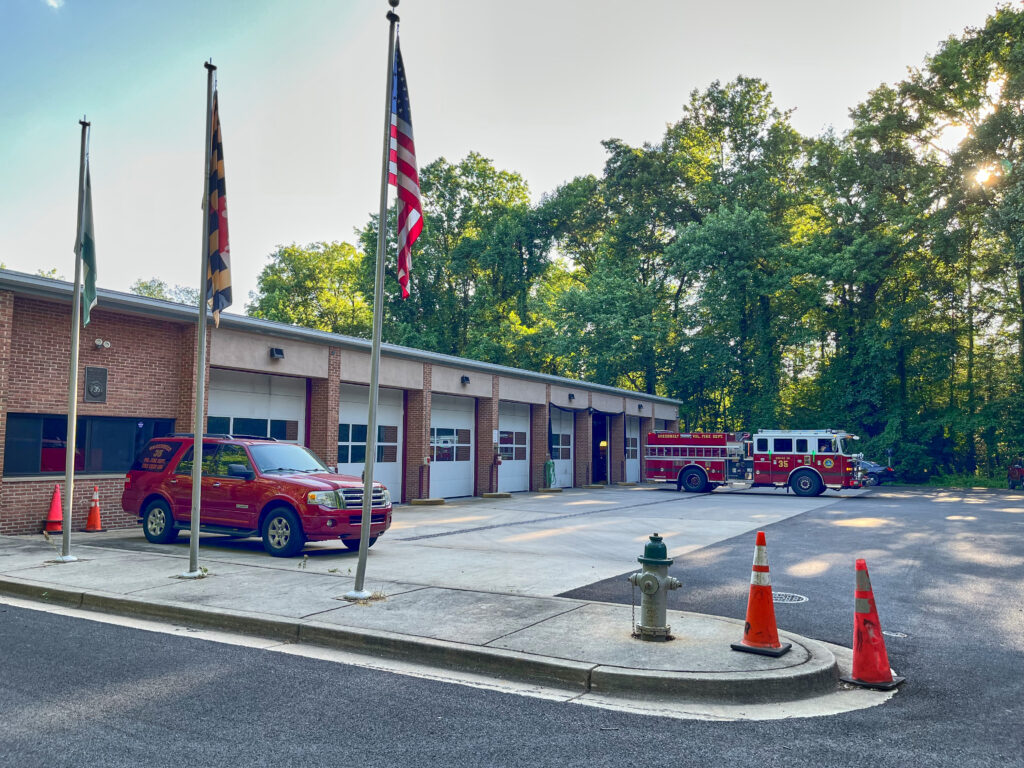 Red-brick single-story building with garages at front and three American flags. A red Fire Department vehicle and a red fire engine are also visible.