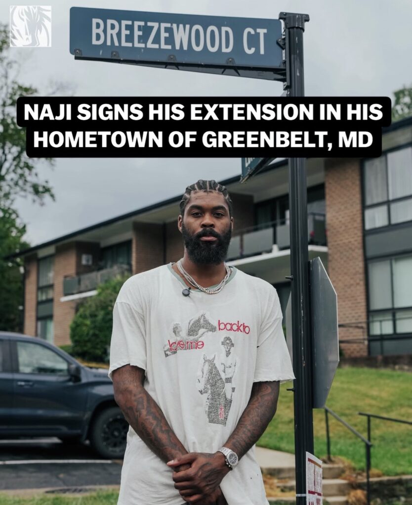 Naji Marshall, a man with brown skin, braided black hair and a black beard, stands below the Breezewood Ct street sign. Behind him is an apartment building. A black banner above his head bears the all-caps caption in white text: NAJI SIGNS HIS EXTENSION IN HIS HOMETOWN OF GREENBELT, MD.