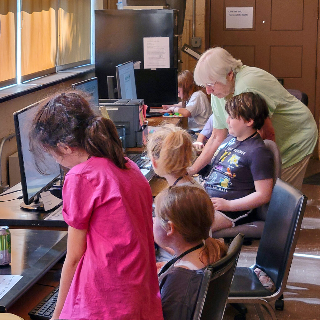 Profile/back view of 5 children sitting at computers at a long table. Cathie Meetre, a woman with short white hair, wearing a green T-shirt and light-colored slacks, stands behind them, leaning over to look at a screen.