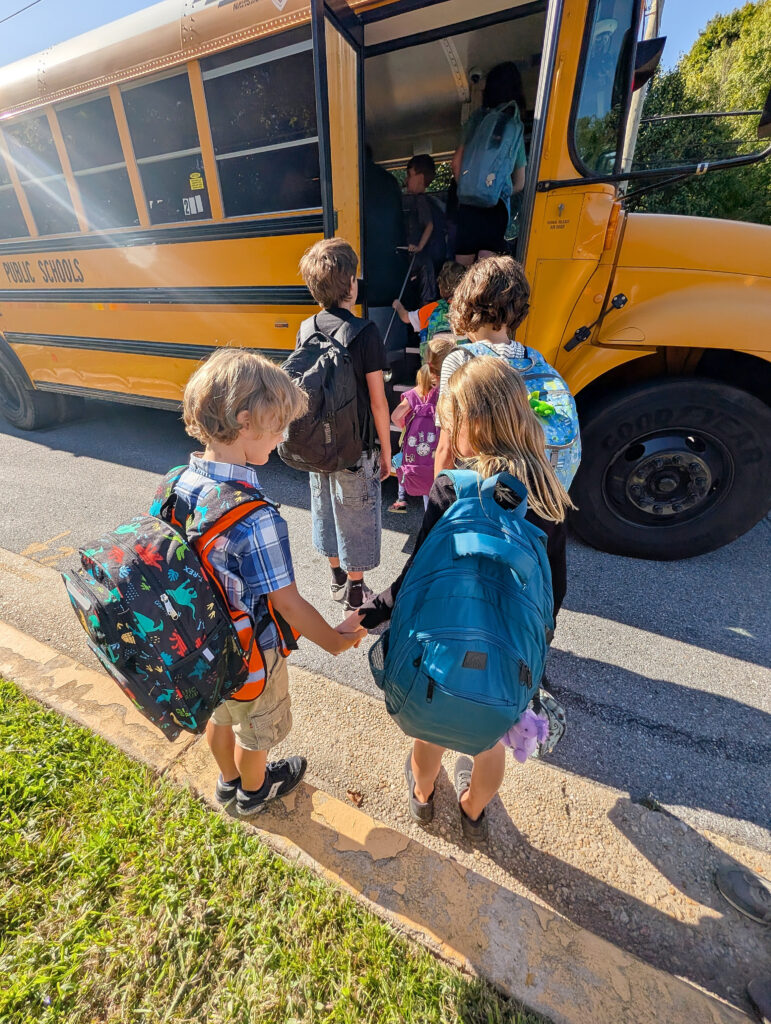 Children wearing backpacks stand in front of the open door of a school bus.