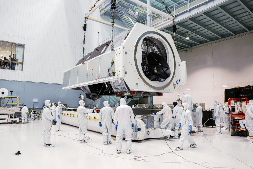 A large white and gray cylindrical structure, on a stand, in a white-walled room. People in full-body white clean-room suits stand around it.