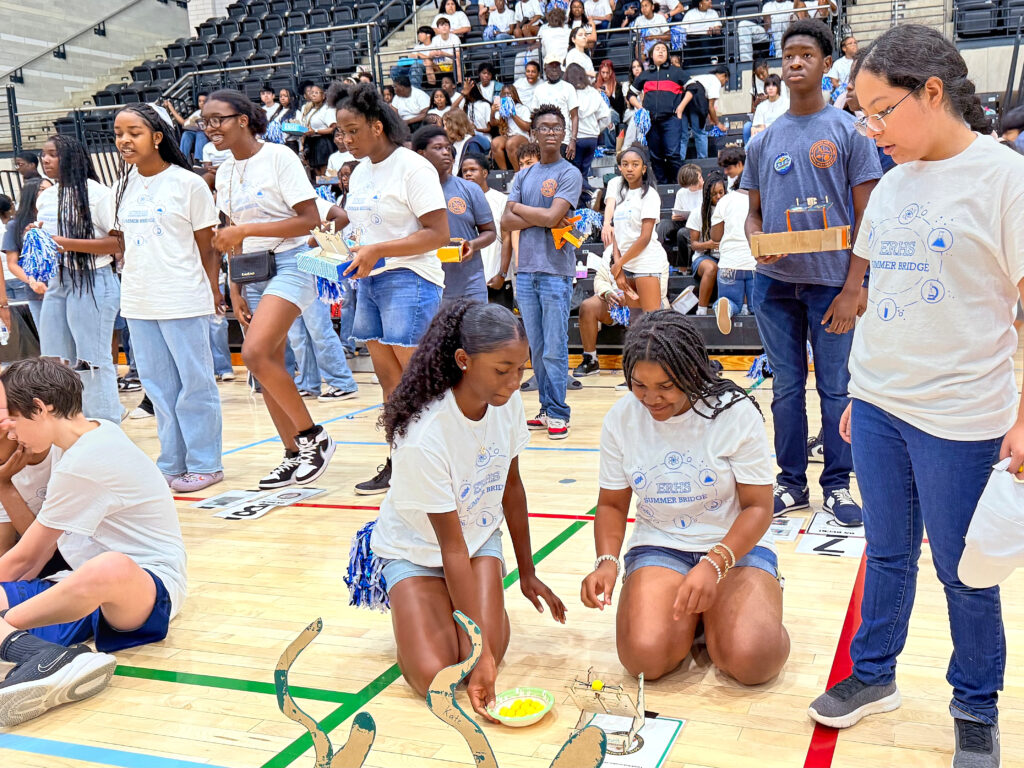 Two high school students kneel on the gymnasium floor, around a catapult project. Other high schoolers stand and sit around and behind them.