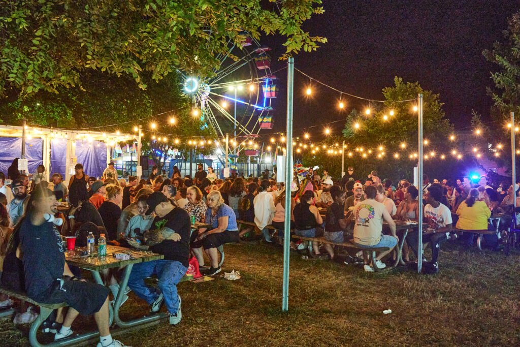 A nighttime photo. People sit at picnic tables while bright yellow globe lights hang overhead. A Ferris wheel is seen in the background.