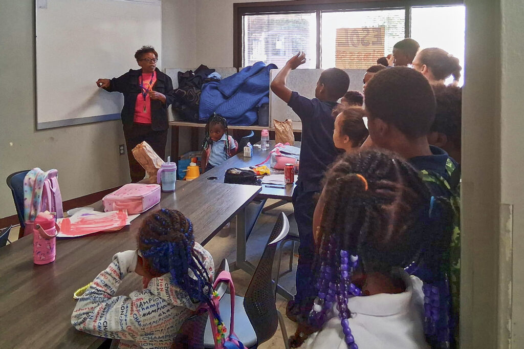 A woman in a pink shirt and black cardigan stands at a whiteboard. Young people are sitting at tables and standing in the back of the room, looking toward her.