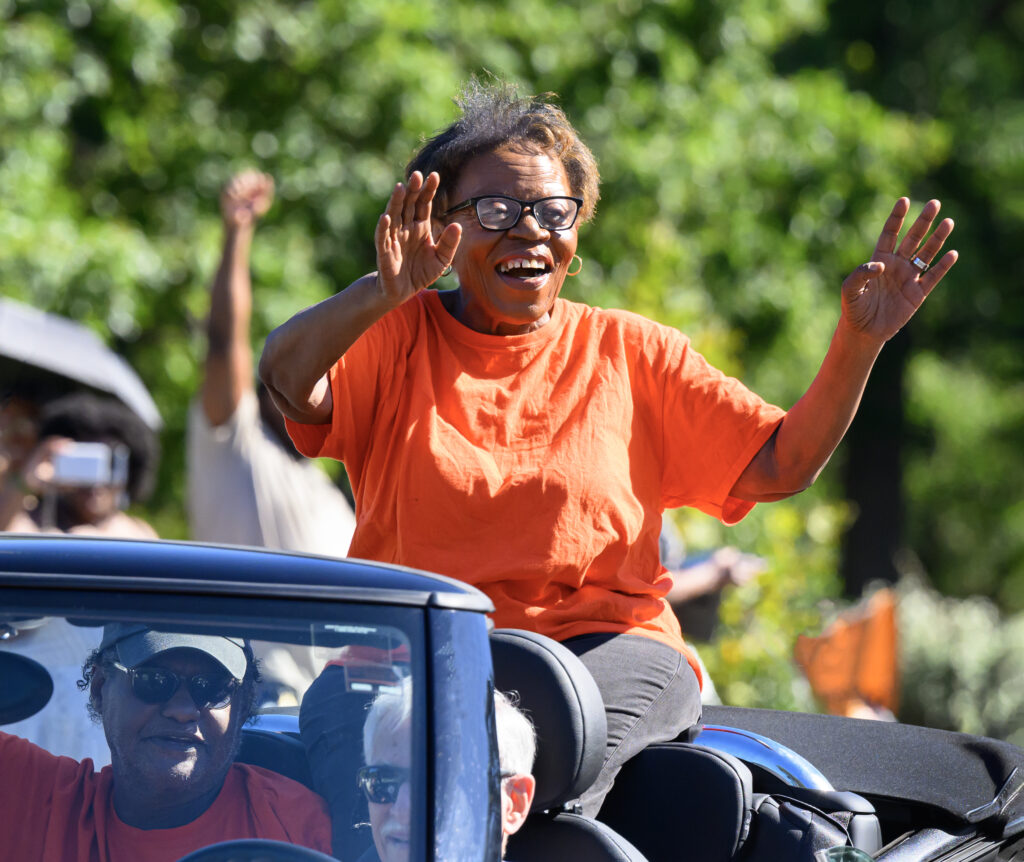 A brown-skinned woman with brown hair, wearing a pink shirt and glasses. She smiles, with hands lifted up, while riding in the back of a convertible car.
