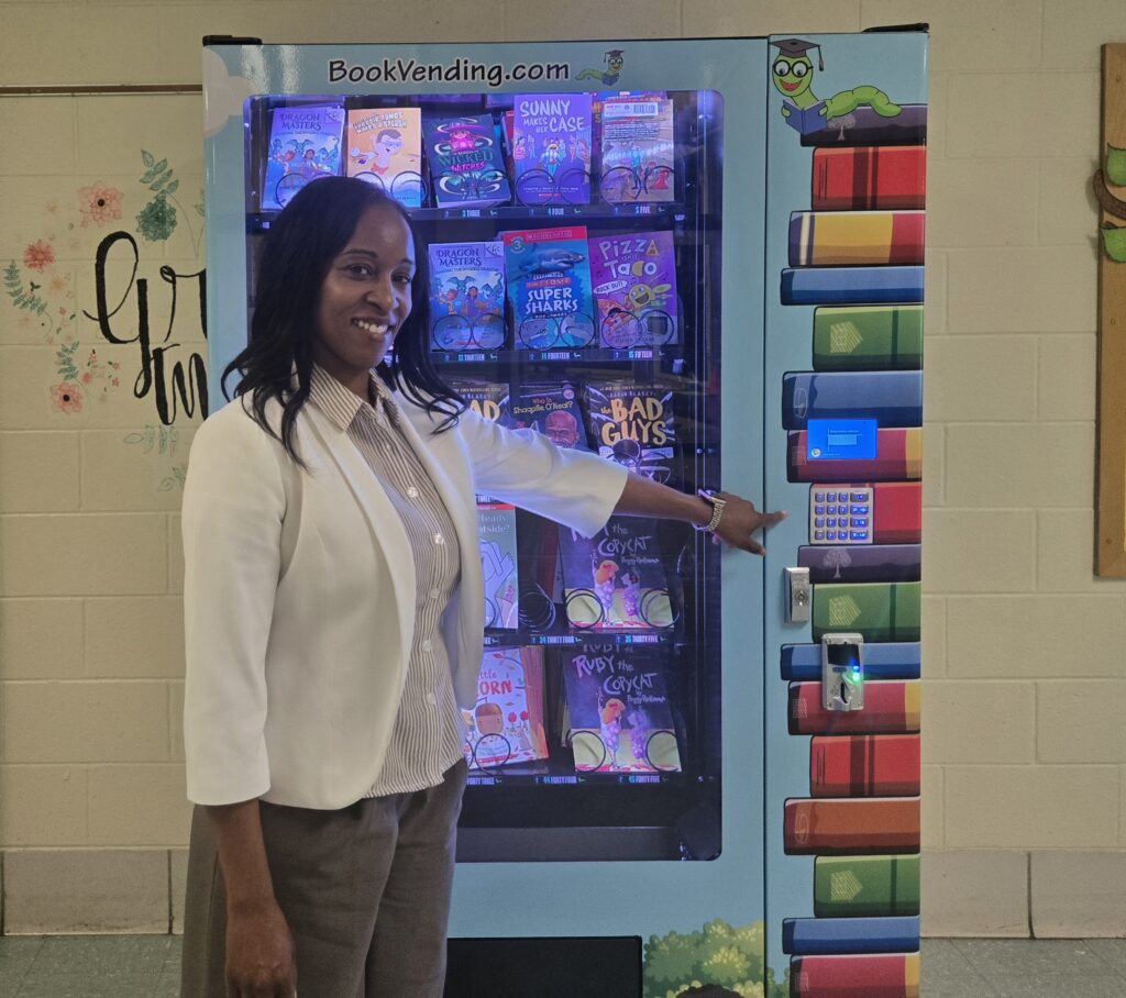 Dorsey, a brown-skinned woman with shoulder-length dark hair, stands in front of the blue vending machine stocked with colorful books. She wears a brown-and-white striped shirt with a white jacket and brown pants. The machine is indoors, against a white-painted wall, with the edge of a mural visible behind it.