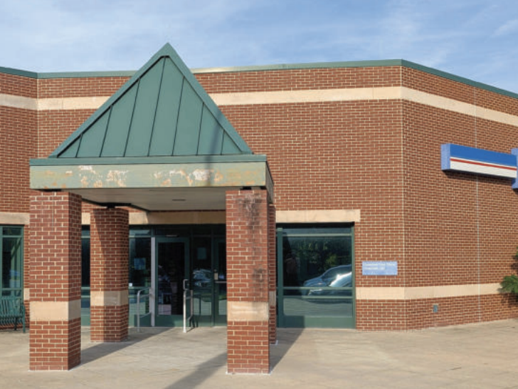 A brown brick building with light beige stripes near the top and bottom. At left is an entryway on matching brick pillars topped by a pointed green roof. At right is a blue, red and white sign.